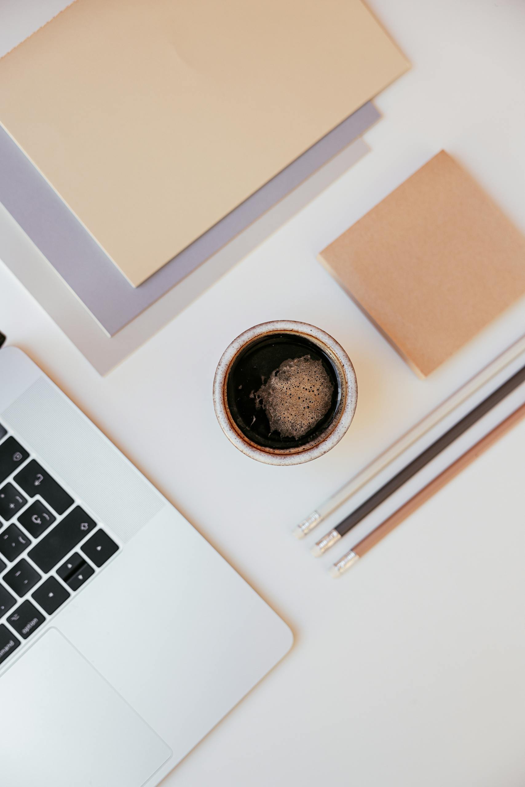 Top-down shot of a workspace with coffee, laptop, notebooks, and pencils on a white surface.